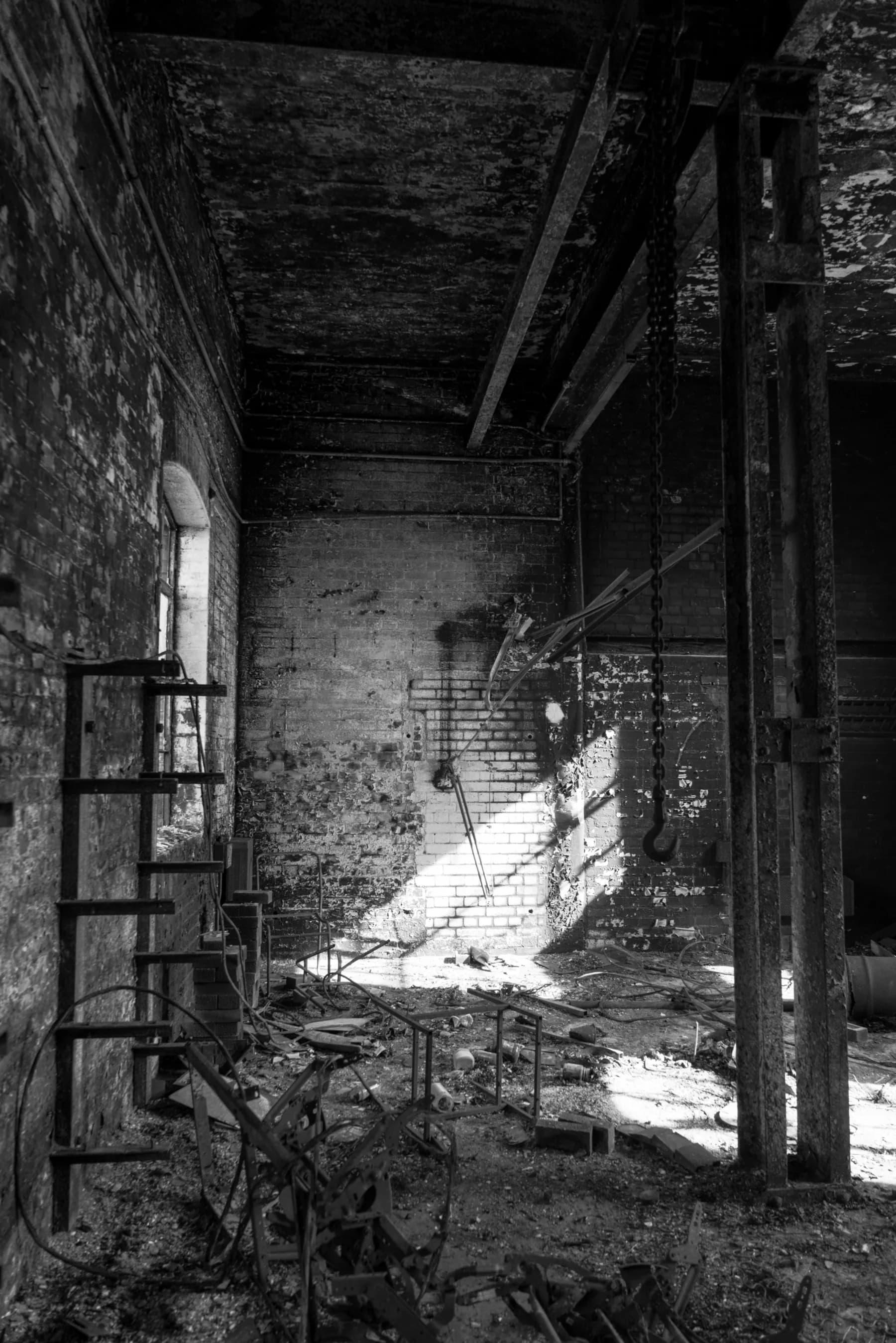 A derelict industrial interior in black and white. A metal ladder on the left, a lifting chain hanging from a timber beam on the right, and a single shaft of afternoon light finding the back brick wall between them.
