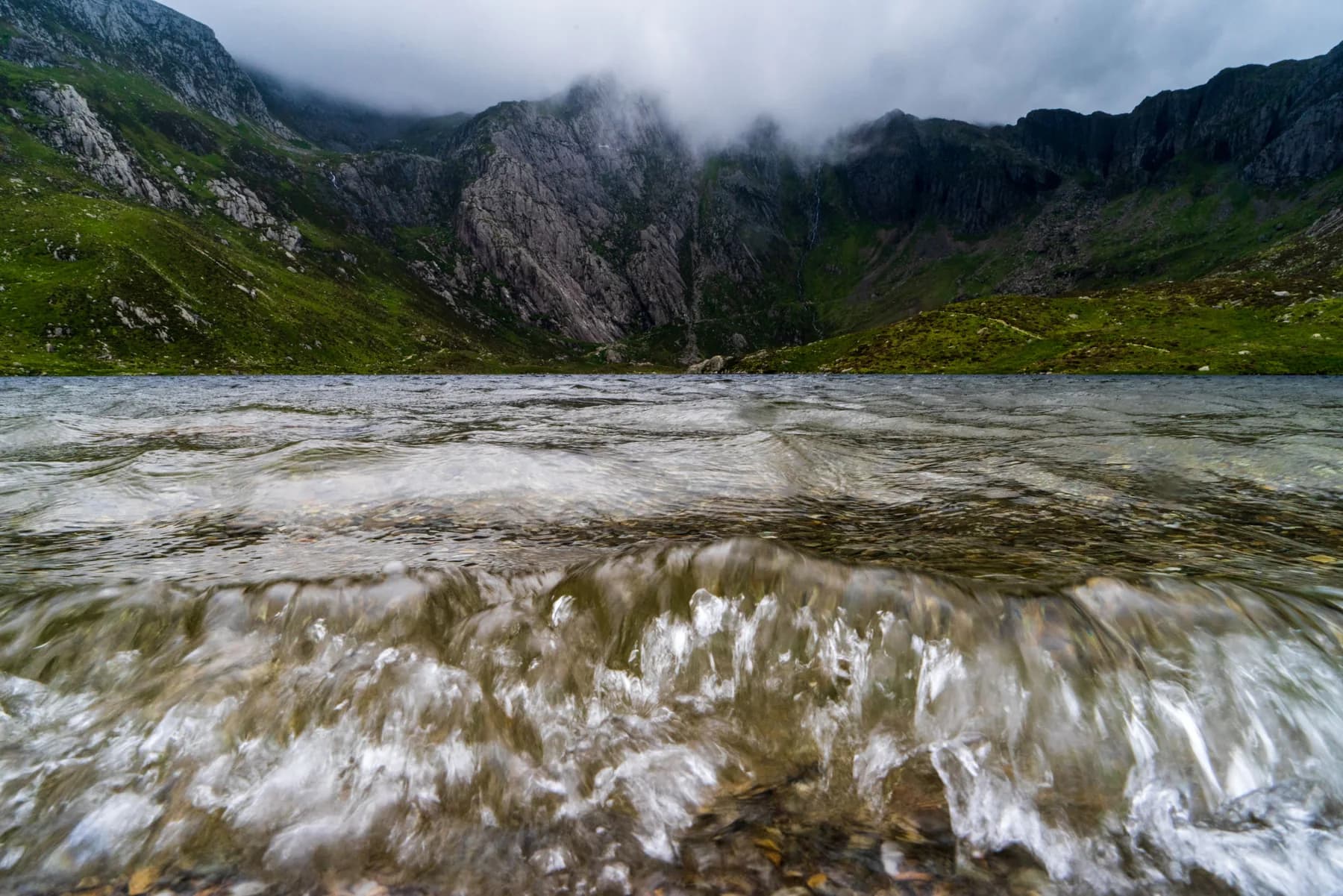 A Welsh mountain cliff-face shrouded in low cloud, rising above a dark lake; churning water and pebbles in the immediate foreground
